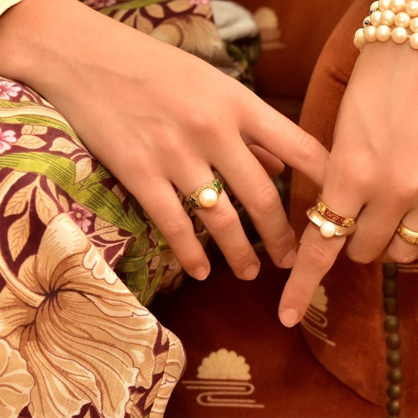 Alt text: Close-up of hands with vintage pearl and enamel rings. One hand wears a bold floral print dress, while the other hand holds the dress. The rings feature pearl solitaires set in 18K gold with intricate guilloche enamel designs in emerald green on the bombé-shaped bands. The cocktail rings have an Art Deco vintage style.