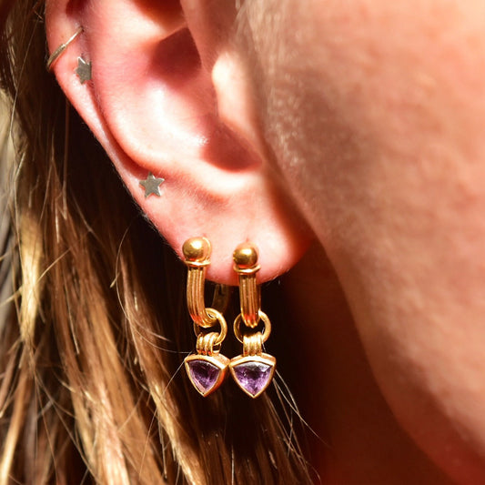 Close-up of an ear wearing small 18K yellow gold hoop earrings with trillion-cut amethyst gemstone charms dangling from the bottom of the hoops.
