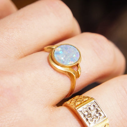 10K yellow gold triplet opal cocktail ring with milgrain bezel and openwork shoulders, featuring an iridescent round opal stone flanked by small diamond accents, shown on a hand model's fingers against a dark background, estate jewelry piece, size 8 1/4 US ring.