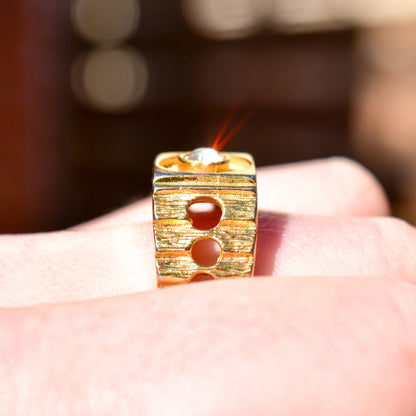 18K gold-plated textured signet ring with circular cutouts, held between thumb and forefinger, blurred background