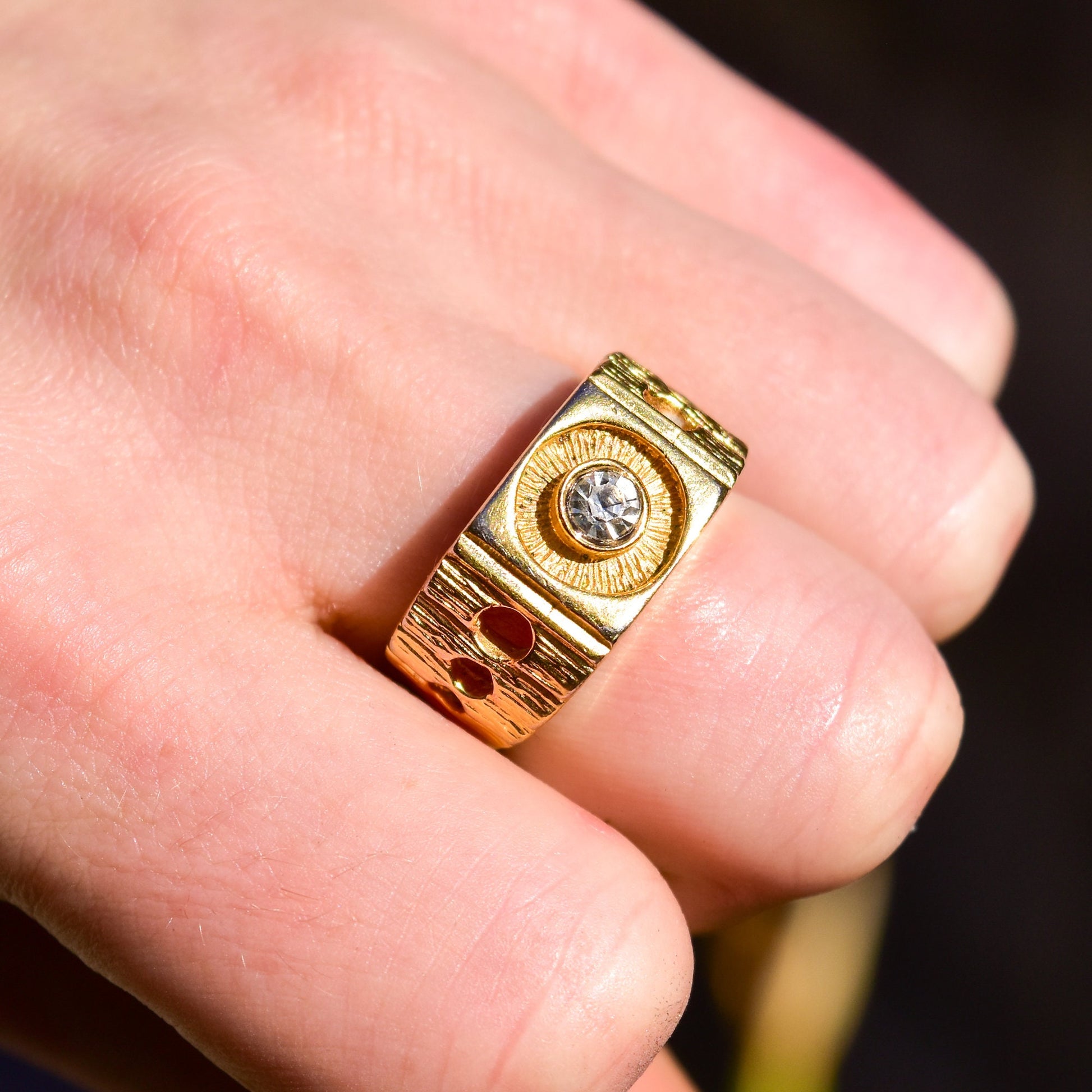 18K gold-plated diamond signet ring with textured chunky design and circle cutouts, shown on a person's finger against a pink background.