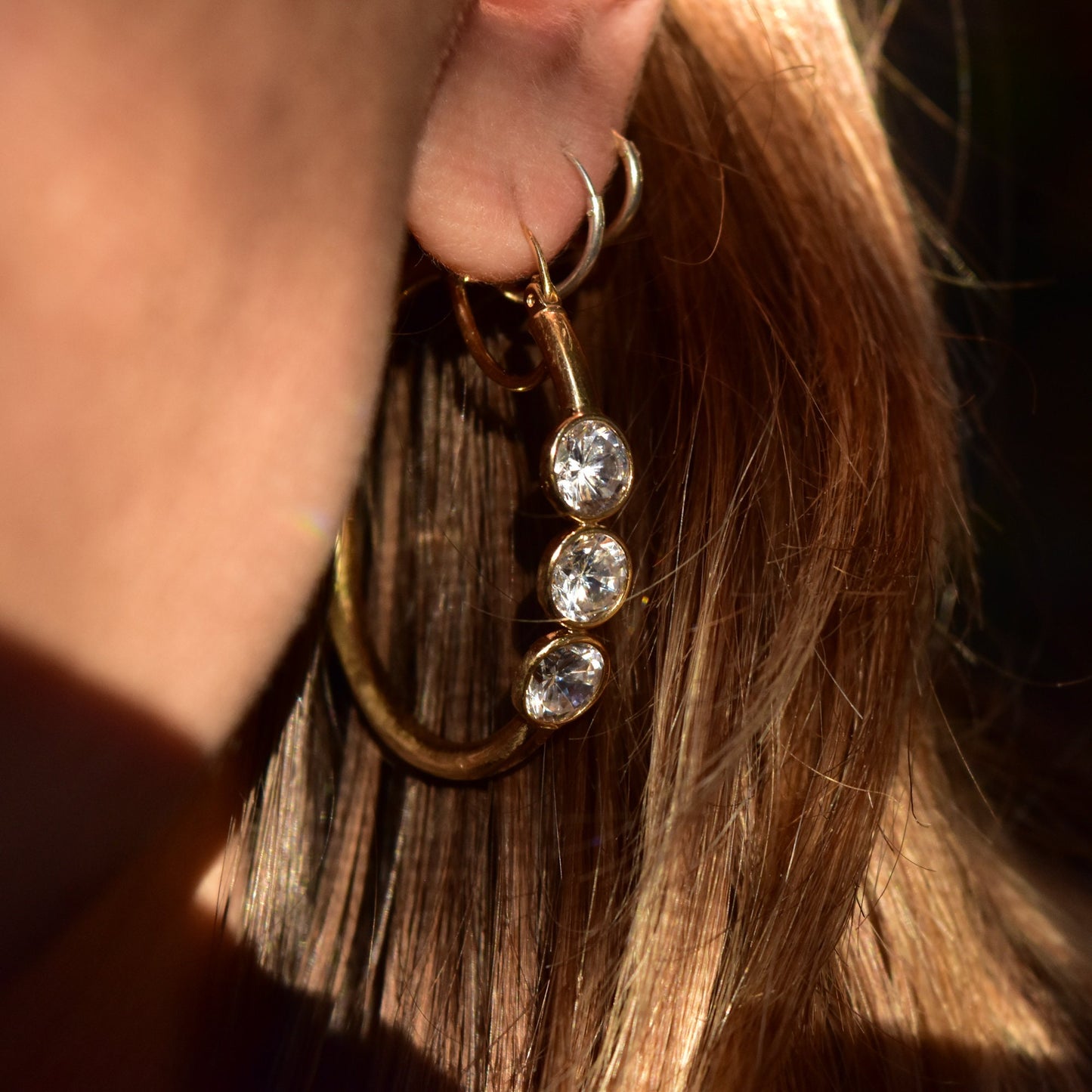 14K yellow gold three-stone cubic zirconia hoop earrings hanging from a woman's ear, with long blonde hair visible