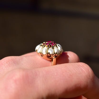 14K yellow gold pearl and ruby cluster ring featuring a domed floral design with white pearls surrounding a center ruby, shown on a finger against a brown background.