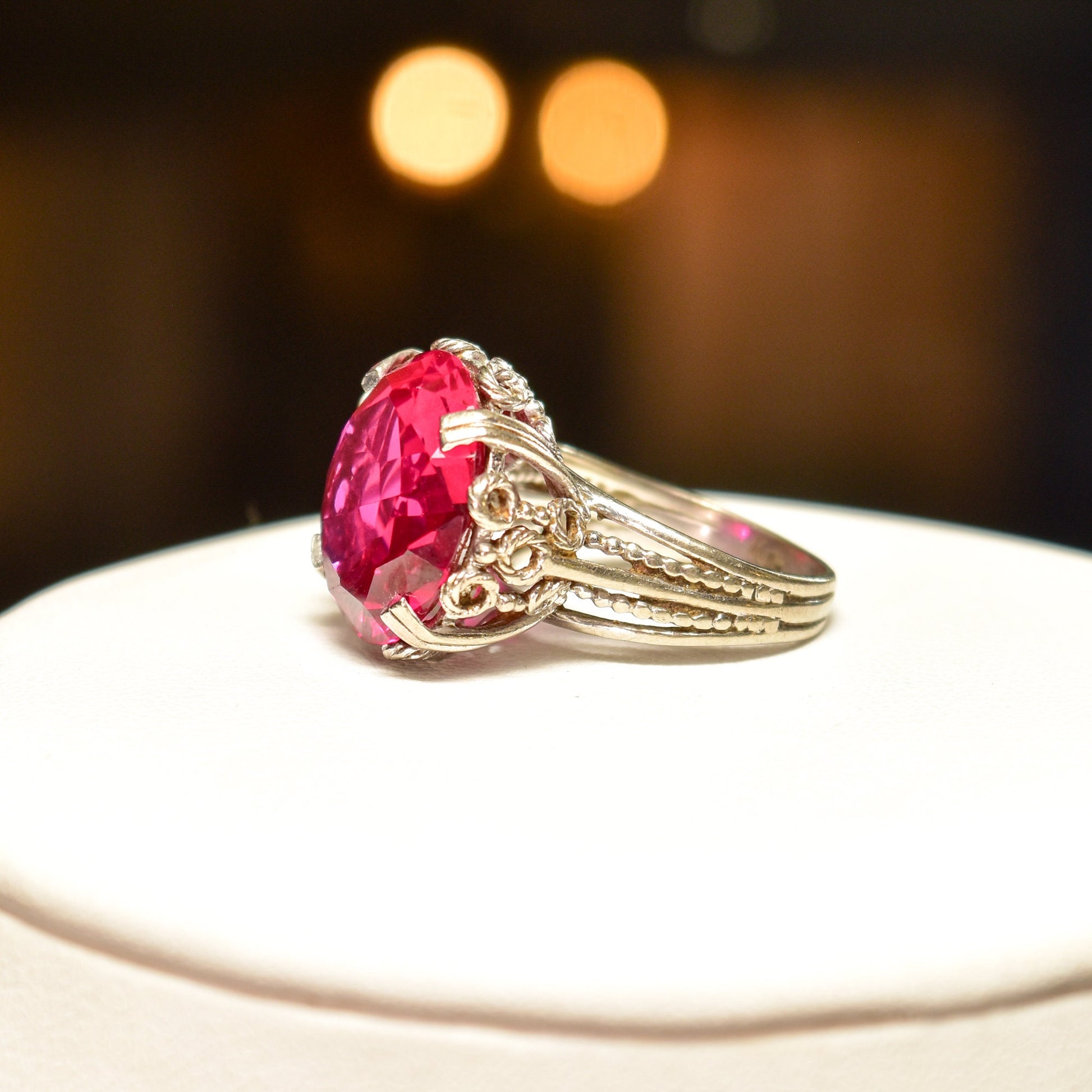 10K white gold Art Deco pink synthetic ruby cocktail ring with ornate filigree setting, size 6 3/4 US, on white background with blurred lights in the background.