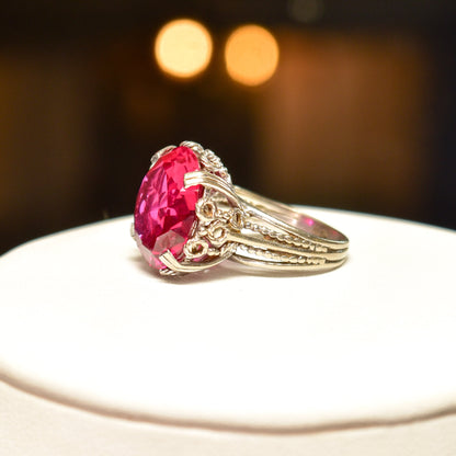 10K white gold Art Deco pink synthetic ruby cocktail ring with ornate filigree setting, size 6 3/4 US, on white background with blurred lights in the background.