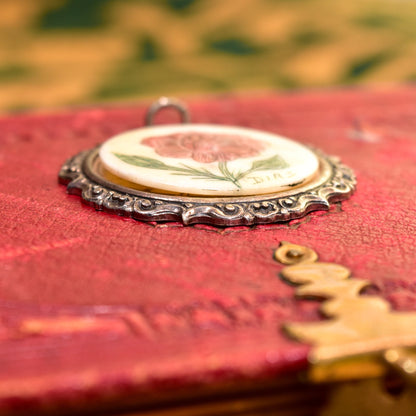 Signed scrimshaw flower pendant in sterling silver with hand-etched and inked bovine bone, set in ornate repousse setting, measuring 44mm in diameter, resting on antique red book cover.