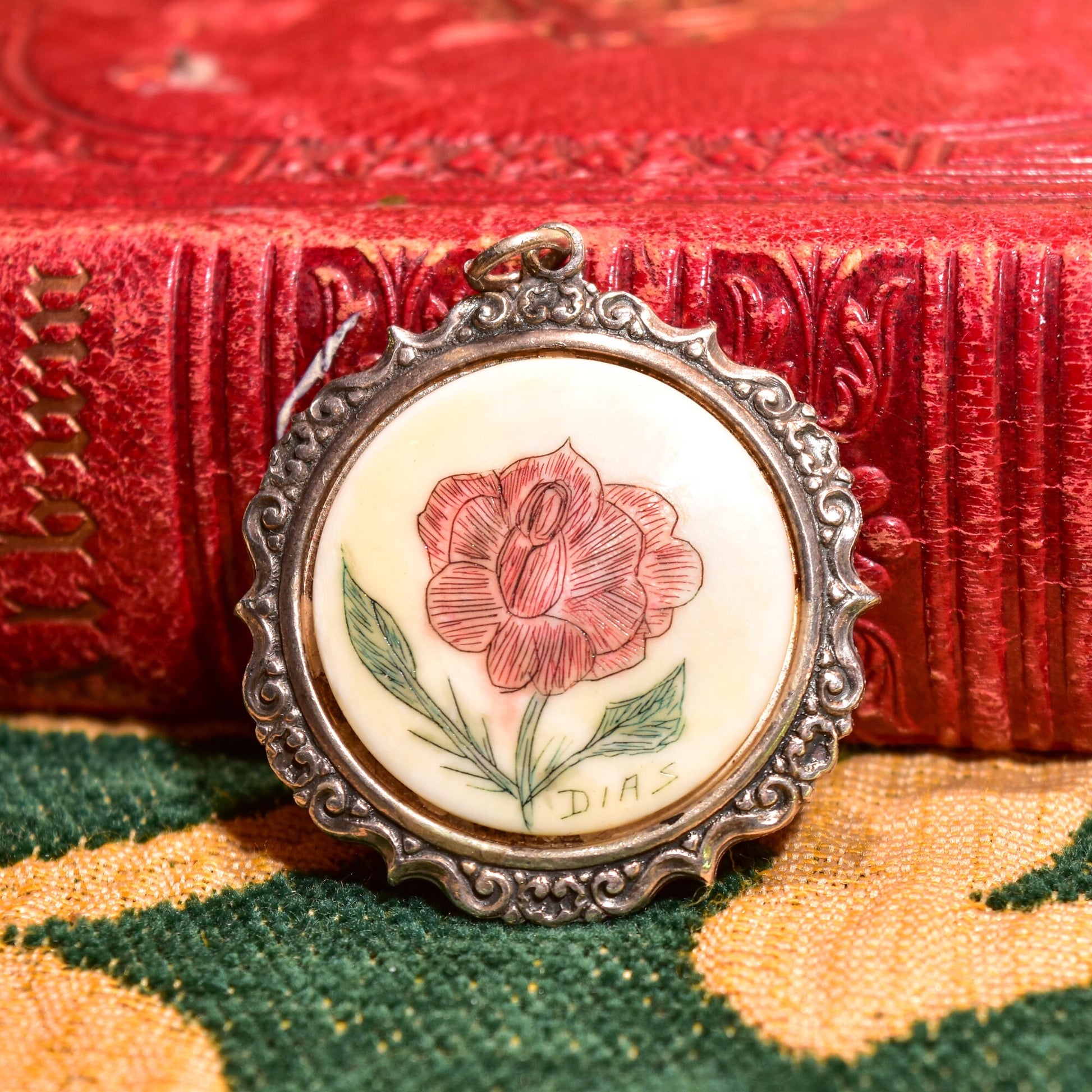 Signed scrimshaw flower pendant in sterling silver setting, featuring a hand-etched and inked red rose design on bovine bone, with an ornate repousse border, measuring 44mm in diameter, displayed on a colorful woven textile background.