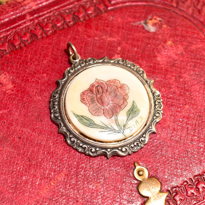 Signed scrimshaw flower pendant in sterling silver setting, hand-etched and inked on bovine bone, featuring a detailed rose bloom design, set in an ornate repousse style frame measuring 44mm in diameter, shown on a textured red fabric background.