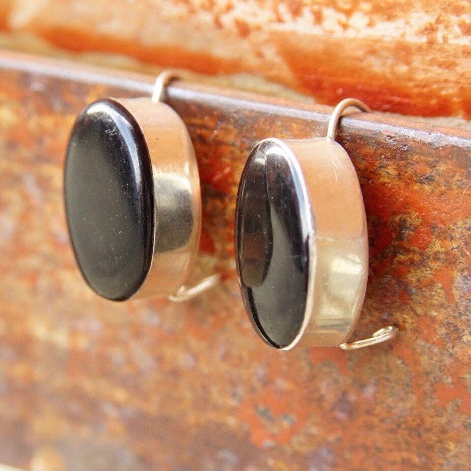 Vintage Taxco modernist sterling silver onyx drop earrings featuring black oval onyx stones set in sleek silver bezels, suspended from silver latch hook wires against a textured rust-colored metal background.