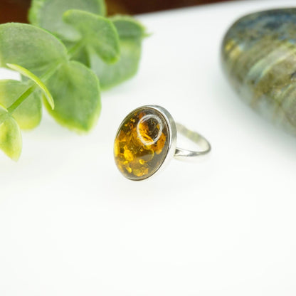 Vintage sterling silver ring featuring an oval amber cabochon with natural inclusions, set in a minimalist design. The ring is shown with green leaves in the background.