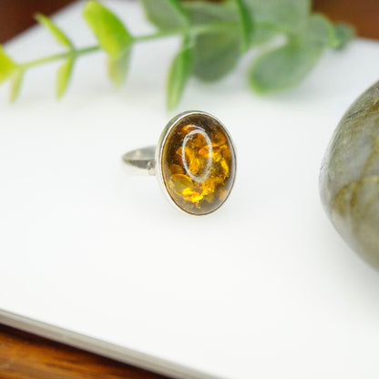 Vintage sterling silver ring with an oval natural amber stone displaying beautiful inclusions, set in a minimalist 925 silver band, photographed on a light background with green foliage.