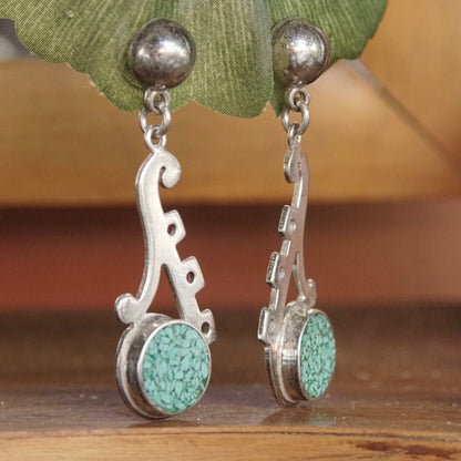 Vintage TAXCO sterling silver earrings featuring mosaic turquoise inlay in an abstract cutout design, dangling from round silver posts against a wooden surface and green leaf background.