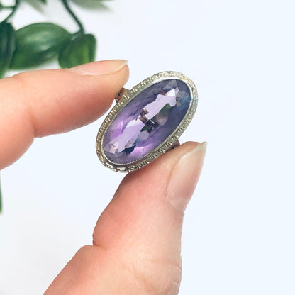 Vintage 18K white gold ring with oval-cut amethyst gemstone held between fingers against white background, showcasing intricate metalwork setting and vibrant purple hue of the amethyst jewelry piece