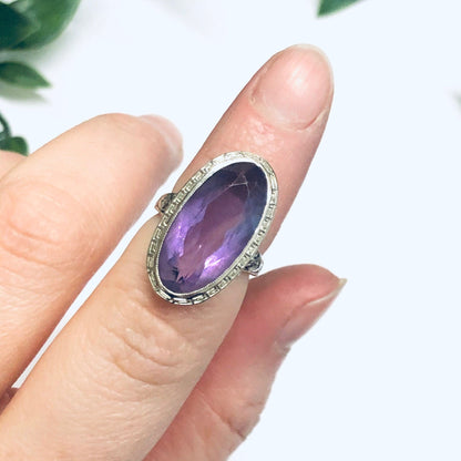 Vintage 18K white gold ring with oval-cut amethyst gemstone held by fingers with green leaves in the background