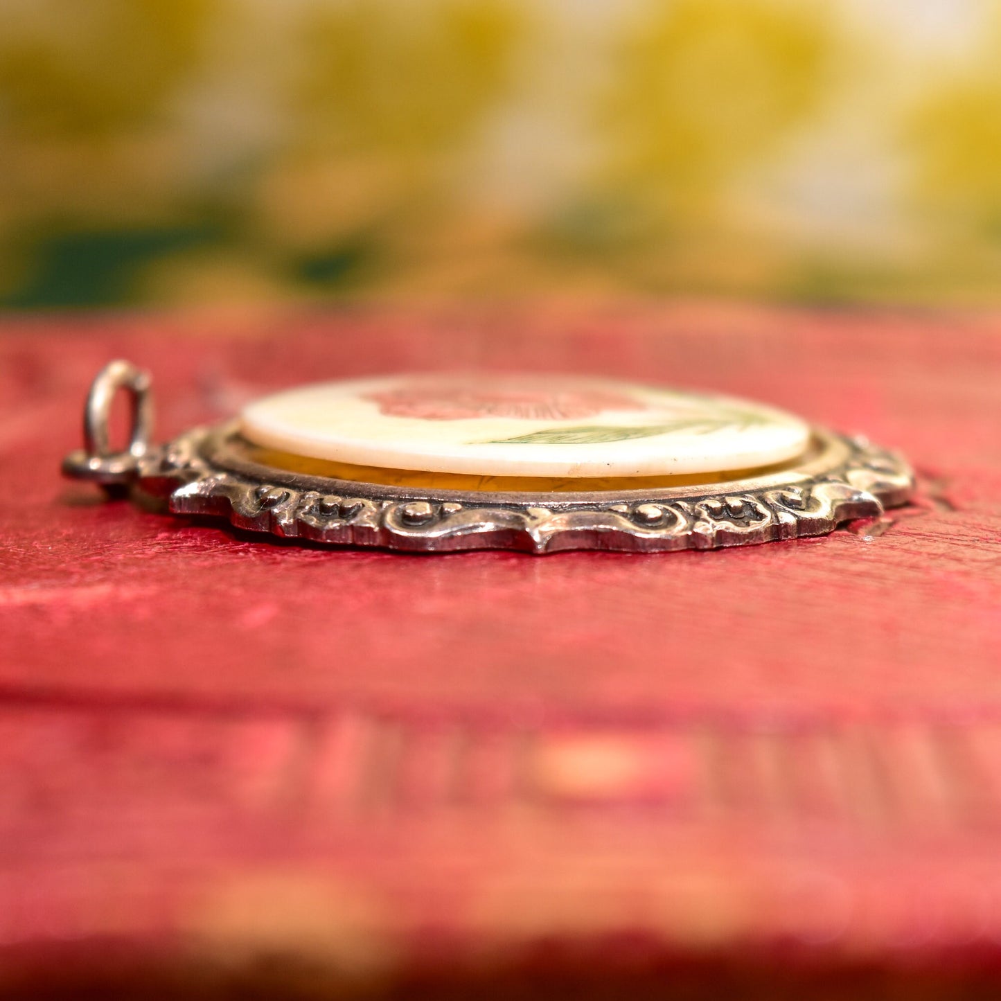 Antique sterling silver pendant with hand-etched floral scrimshaw on bovine bone, set in an ornate repousse frame, measuring 44mm in diameter, resting on a red textured surface with a blurred yellow and green background.