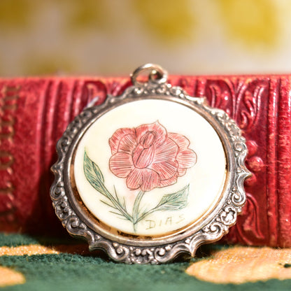 Hand-etched and inked scrimshaw pendant featuring a red rose flower design, set in an ornate sterling silver frame against a textured red velvet background.