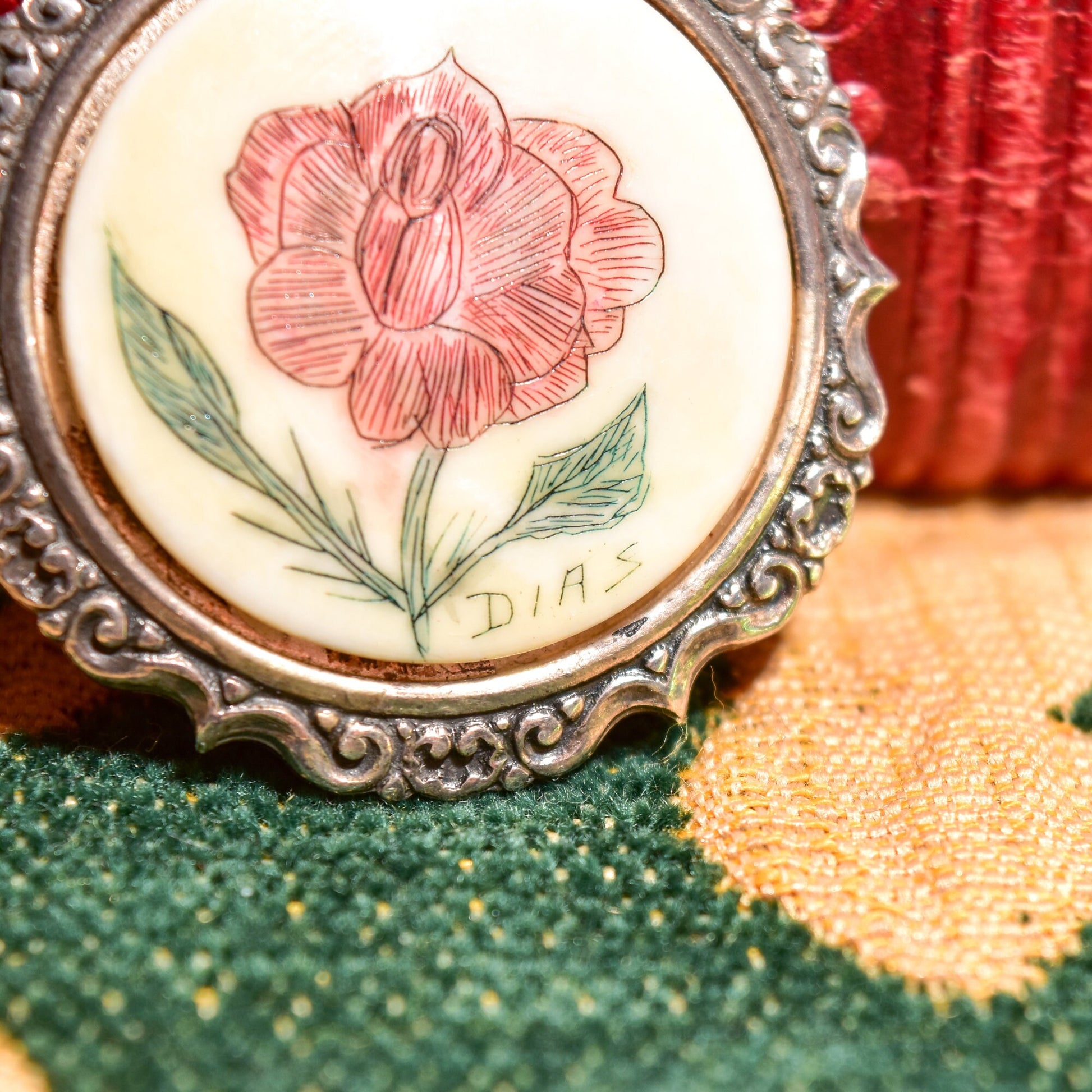 Scrimshaw pendant featuring a hand-etched red flower on bovine bone, set in an ornate sterling silver repousse frame, on green textured fabric