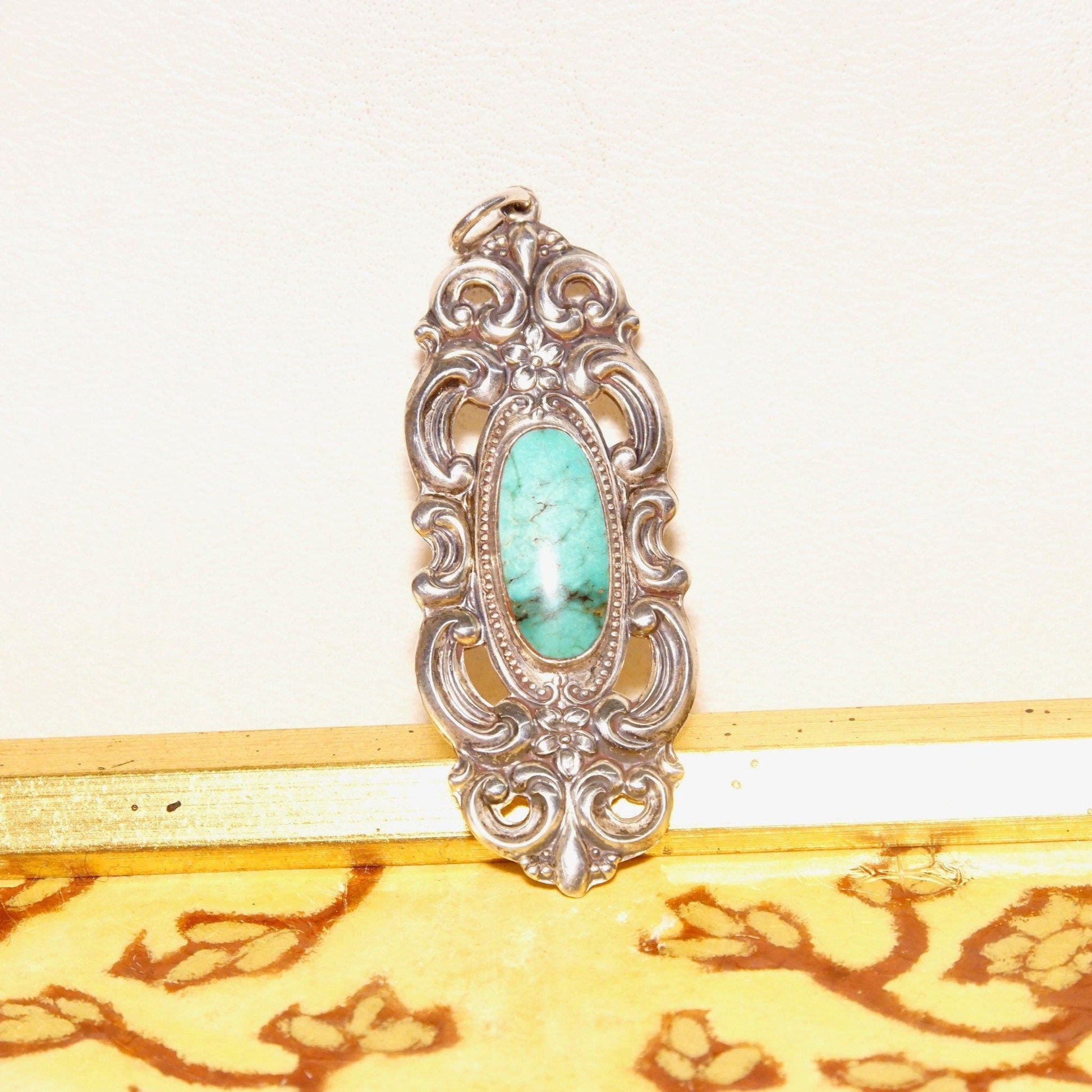 Vintage Towle sterling silver pendant with ornate embossed repousse designs, featuring a turquoise cabochon at the center. The pendant has an art nouveau style with intricate floral motifs and scrollwork, measuring 2 3/4 inches in length.