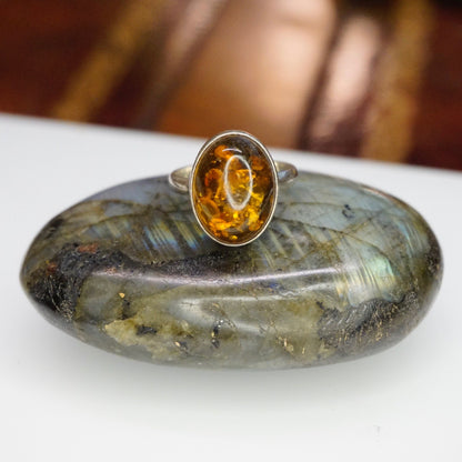 Vintage sterling silver ring with an oval amber stone displaying unique natural inclusions, set in a minimalist 925 silver setting on a textured gray background.