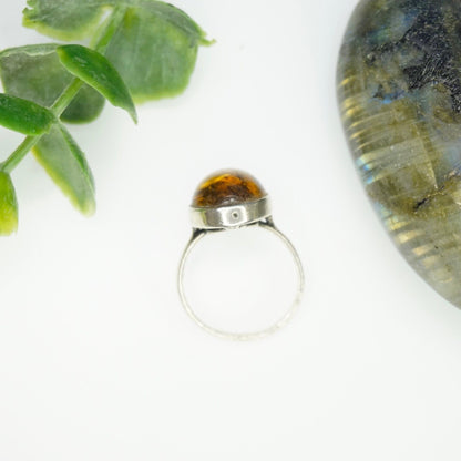 Vintage sterling silver ring with an oval amber stone displaying natural inclusions, set in a minimalist band, against a white background with green leaves.