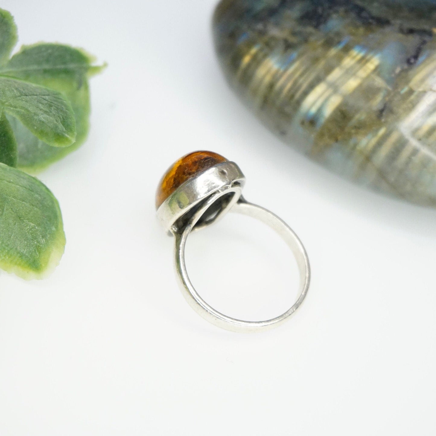 Vintage sterling silver ring with a beautiful oval amber stone featuring natural inclusions, presented on a white background with green leaves and a decorative shell in soft focus.