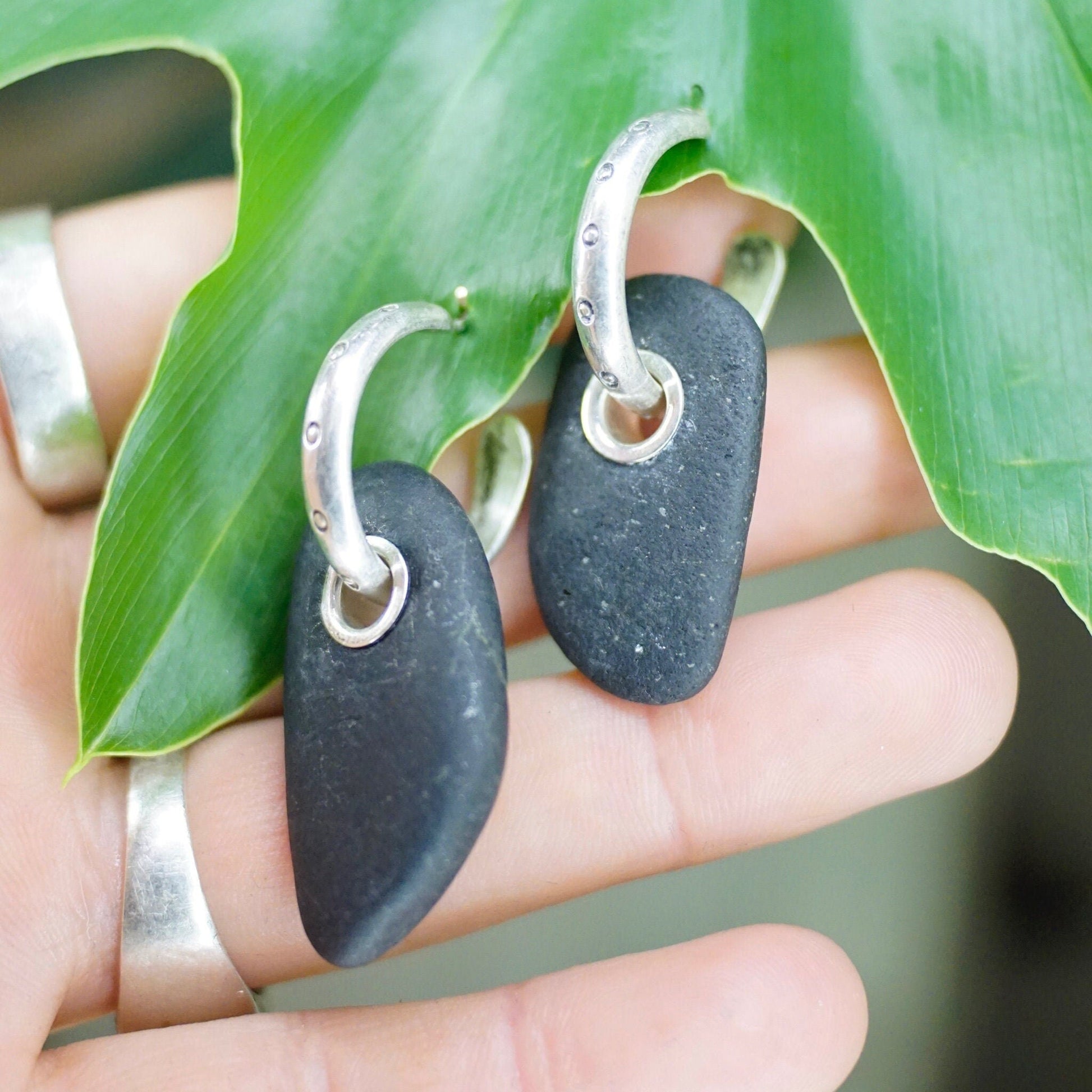 Vintage sterling silver hoop earrings with black stone charms on hand holding green leaf, artisan handmade 925 silver statement earrings with threaded rock pendants.