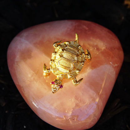 Vintage 14K yellow gold turtle brooch featuring ruby eyes, a detailed shell design, and small ruby accents on the feet, displayed on a pink crystal rock against a black background.