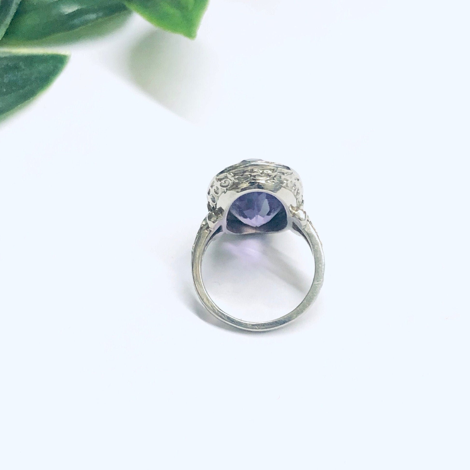 Vintage 18K white gold ring featuring a round amethyst gemstone set in an intricate design. The ring is displayed with green leaves in the background, showcasing the beauty of the purple amethyst and elegant white gold setting. Perfect as an engagement ring or statement piece of 18K jewelry.