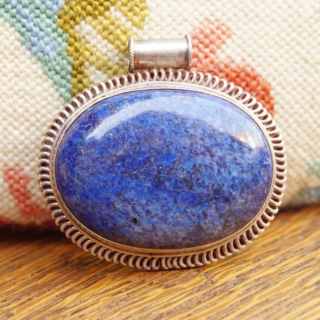 Vintage modernist sterling silver pendant with large marbled blue lapis lazuli gemstone, measuring 2 1/2 inches long by 2 3/4 inches wide, against a colorful woven textile background.