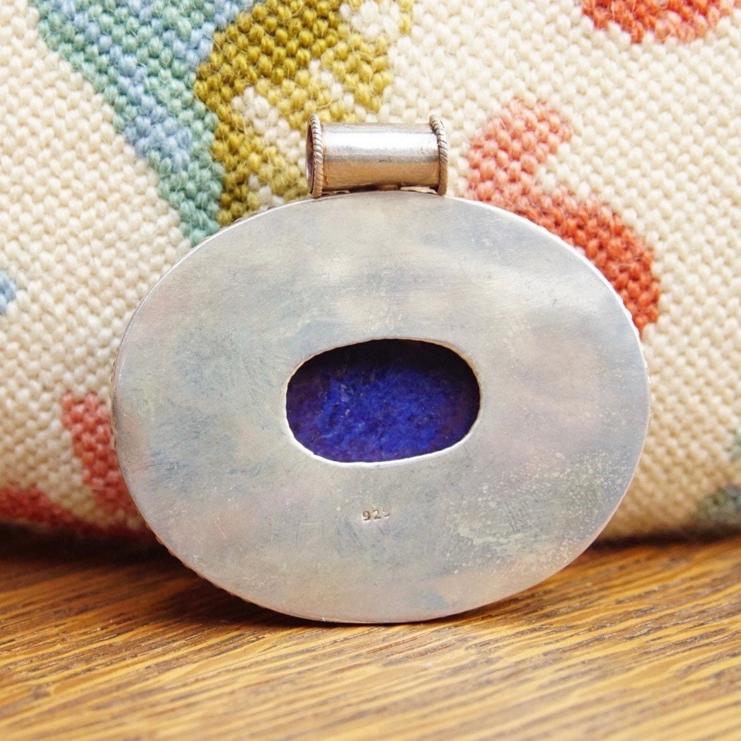 Vintage modernist sterling silver pendant featuring a large, marbled blue lapis lazuli gemstone, measuring approximately 2 1/2 inches long by 2 3/4 inches wide, displayed on a colorful woven textile background.