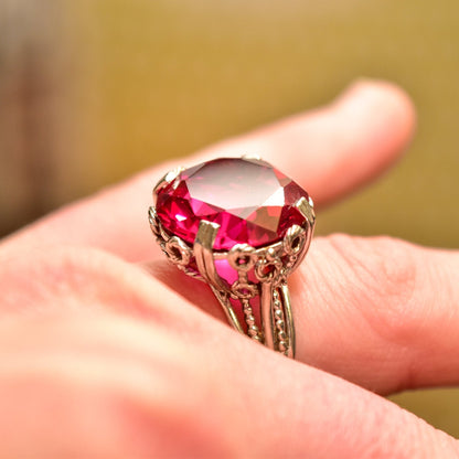 10K white gold ornate filigree cocktail ring with large round pink synthetic ruby gemstone, Art Deco style, held on finger, size 6 3/4 US.