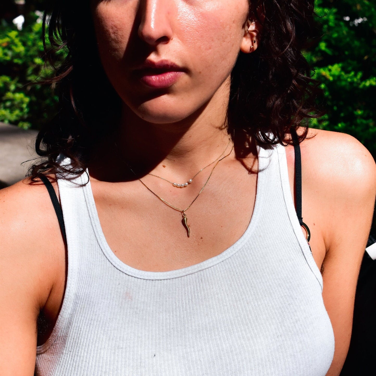 Close-up of a woman wearing a small gold Italian horn charm necklace against her skin, with green foliage visible in the background on a sunny day.