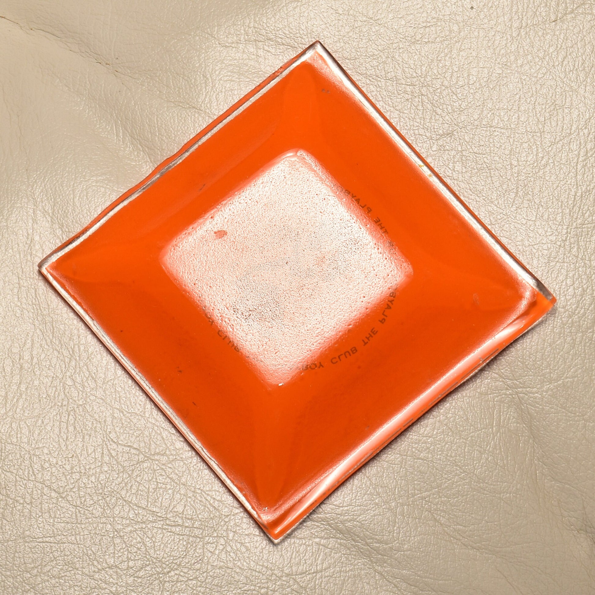 1960's The Playboy Club orange glass ashtray with Leroy Neiman Femlin illustration, small vintage trinket dish measuring 4.75 inches square, against textured background