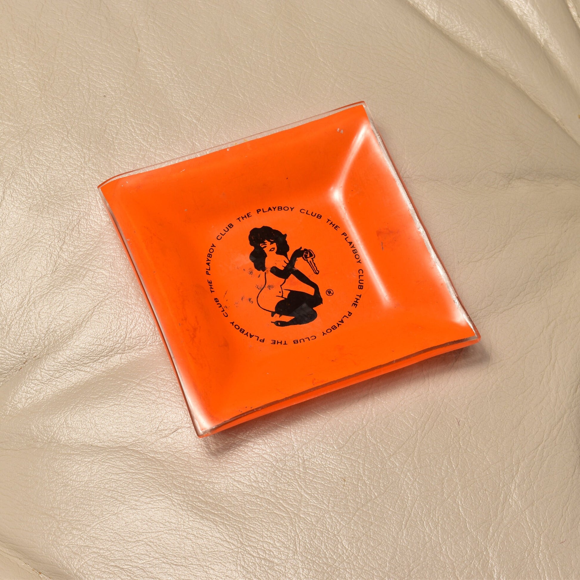 Orange glass Playboy Club ashtray featuring a silhouette illustration of a femlin character by Leroy Neiman on a white cloth background.