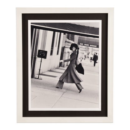 Vintage black and white photograph of a fashionable woman in a hat and long coat walking on a city sidewalk near storefronts, presented as framed wall art.