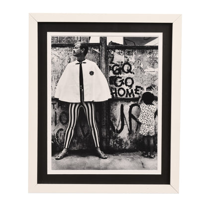 Black and white framed photograph depicting a stylishly dressed person standing in front of a graffiti-covered wall, wearing striped pants and a white cape. The wall behind them has "CQ GO HOME" written on it in graffiti lettering.