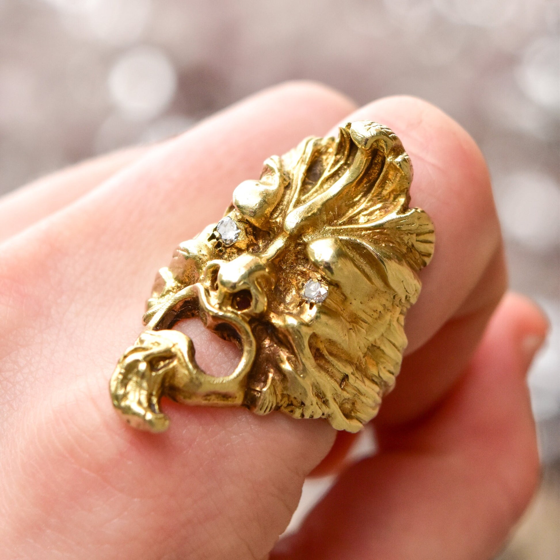 Gold sculpted face ring with diamond eyes held in a hand, close-up view showing intricate detailing of the textured mask design with sparkling diamond accents.