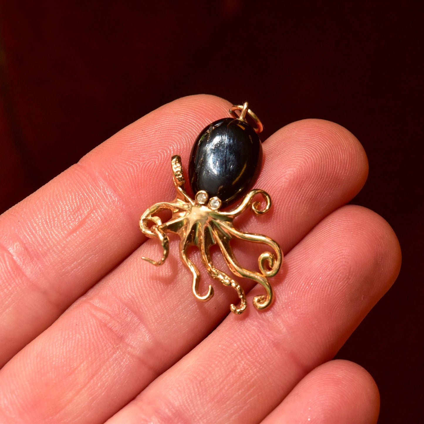 10K yellow gold 3D octopus pendant with black coral cabochon body and diamond eye, held in hand against dark red background, showing intricate tentacle details of the aquatic animal-inspired jewelry piece.
