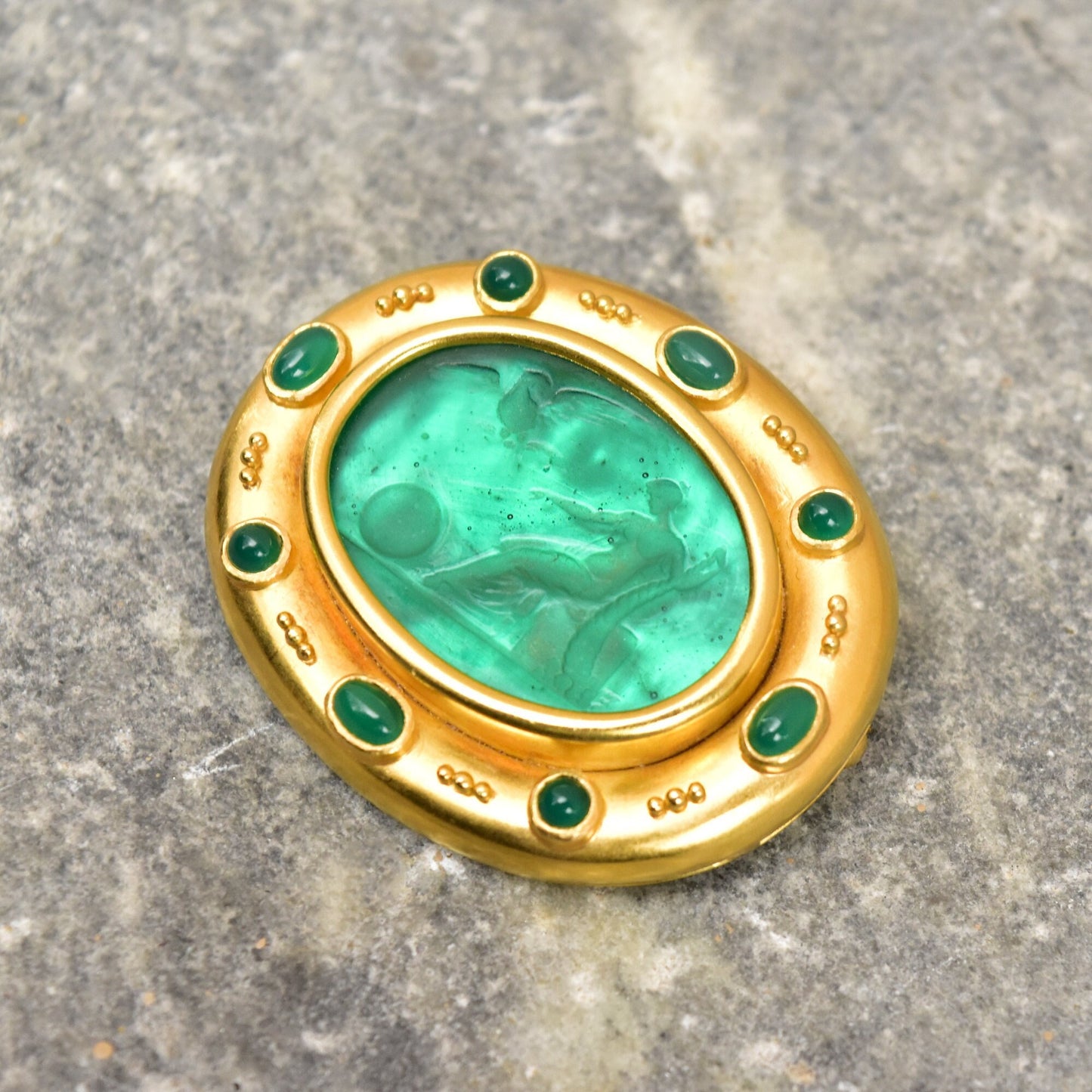 18K gold brooch pendant featuring an oval Venetian glass intaglio in emerald green, accented with small round emerald cabochons, with a mother of pearl backing, measuring approximately 2 inches wide, by Elizabeth Locke.