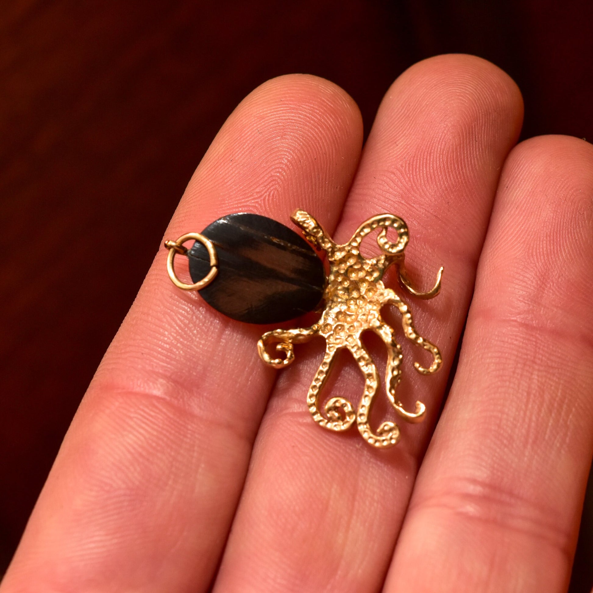 10K yellow gold octopus pendant with black coral center and diamond accents, held in palm of hand against deep red background