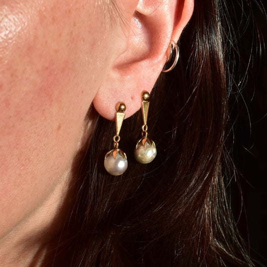 18K yellow gold pierced dangle earrings featuring baroque freshwater pearls, shown on a woman's ear with dark hair, close-up view.