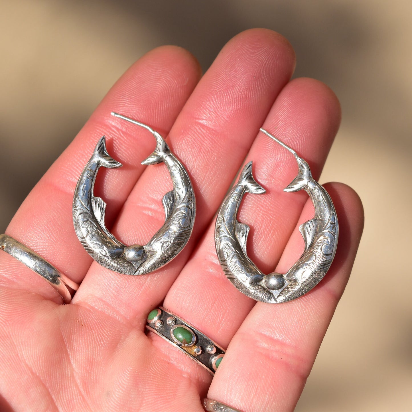 Engraved sterling silver fish hoop earrings in hand, showing mirrored etched fish design on oval pierced hoops measuring 30mm in diameter.