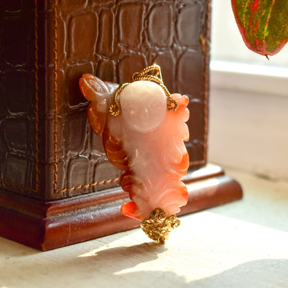 Carved red jade Buddha pendant with yellow gold wire wrapping, displayed on a wooden stand near a window with sunlight streaming in.