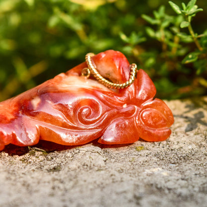 Red jade Buddha pendant with intricate carvings and yellow gold wire wrapping, laying on textured stone surface with green foliage in background.