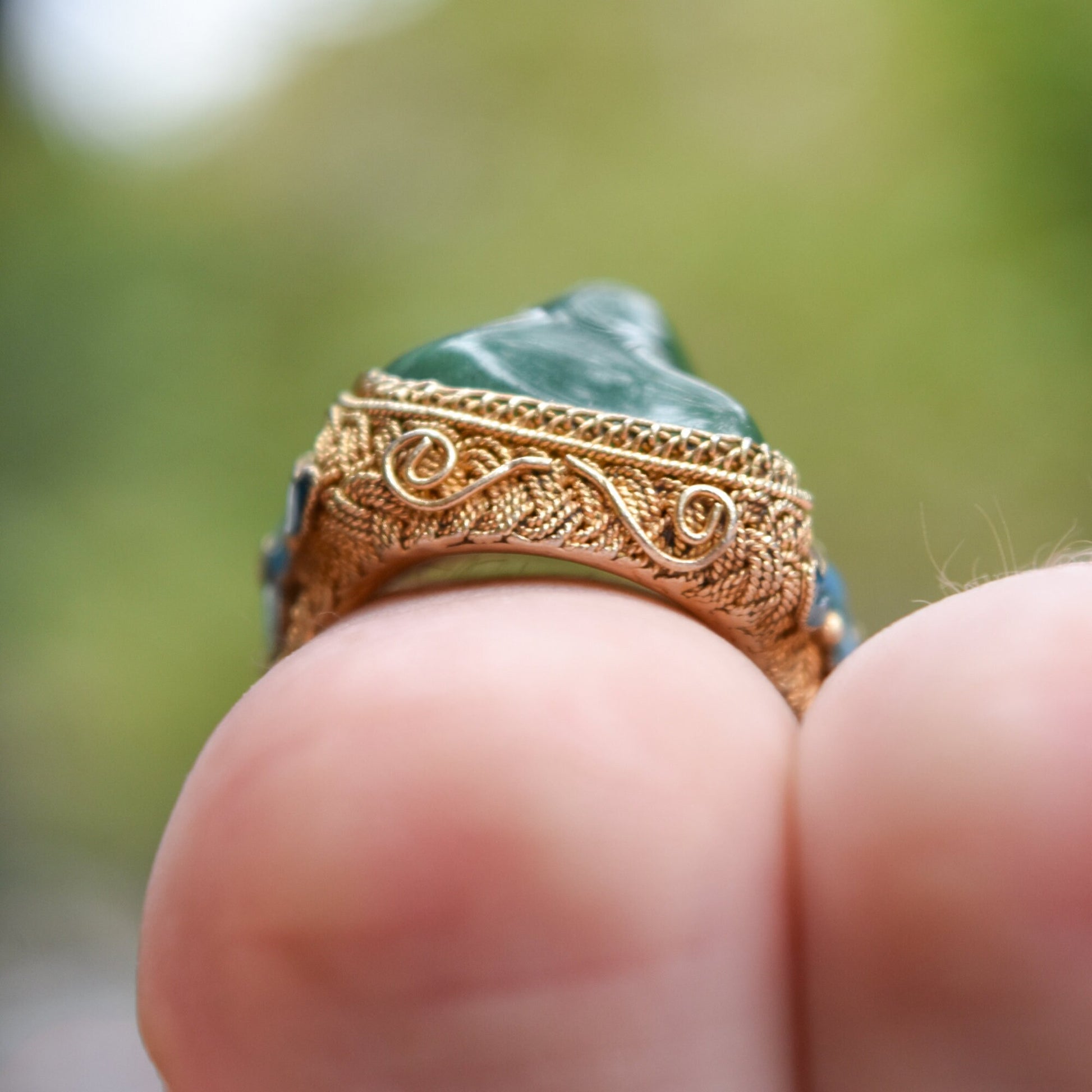 Vermeil silver adjustable ring with intricately carved floral motifs, green jade gemstone and Chinese enamel design, shown on fingers against blurred green background.