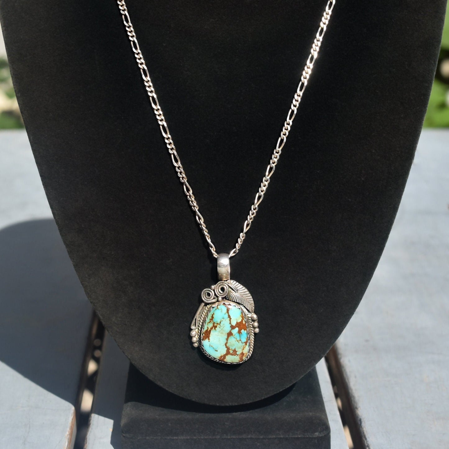 Vintage Native American turquoise pendant necklace crafted in sterling silver, featuring intricate leaf and swirl designs. The pendant measures approximately 1.75 inches in length and showcases a vibrant turquoise stone at the center. An example of traditional Navajo jewelry craftsmanship.