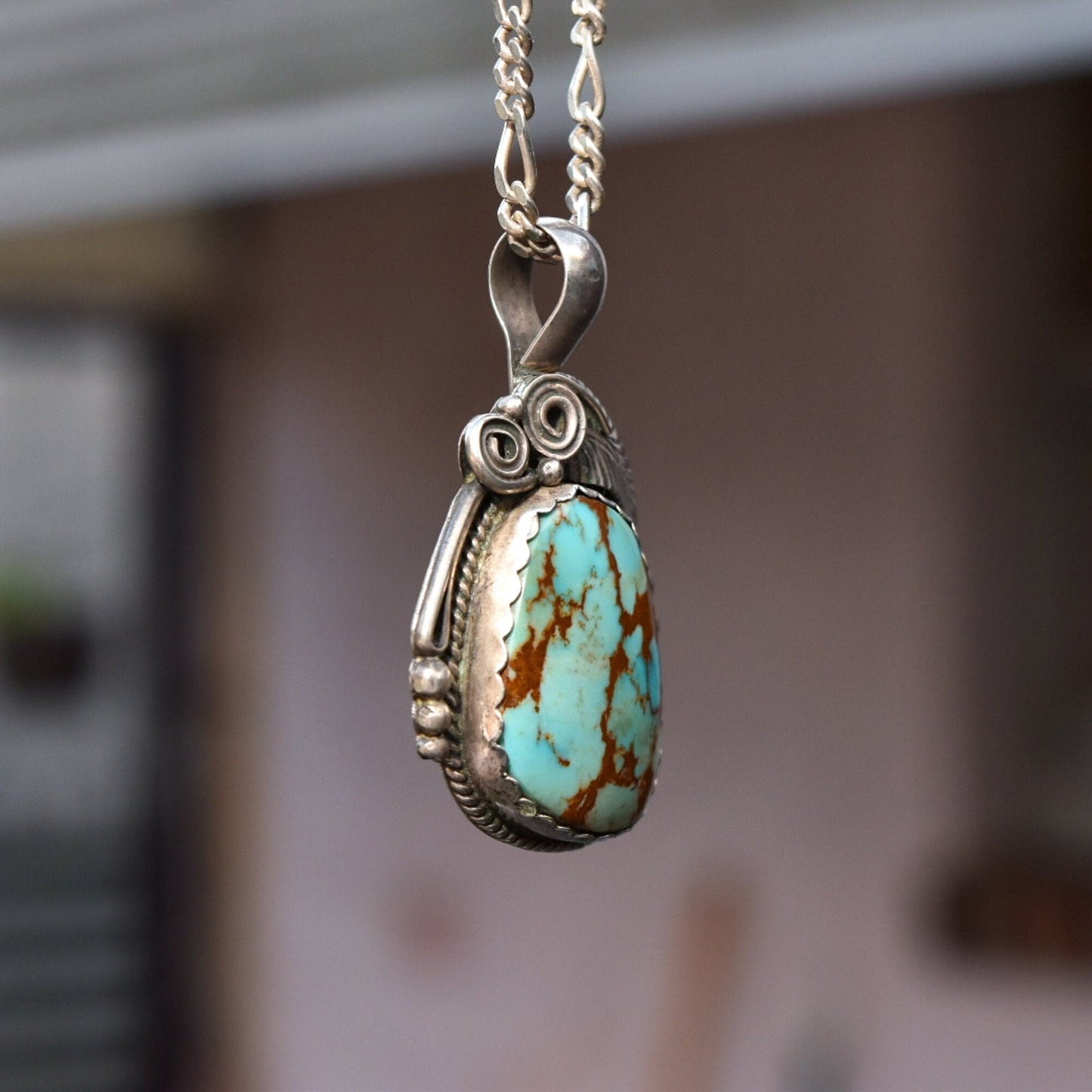 Navajo sterling silver pendant necklace featuring a teardrop-shaped turquoise stone with brown matrix, accented by twisted wire and leaf motifs. The pendant hangs from a silver chain against a blurred background.