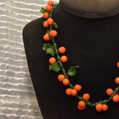 Orange glass berry necklace with green leaves on black mannequin against wavy textured background