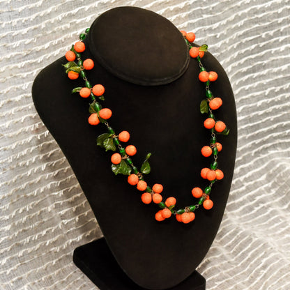 Venetian glass orange berry necklace with green leaves on black bust display against textured white background