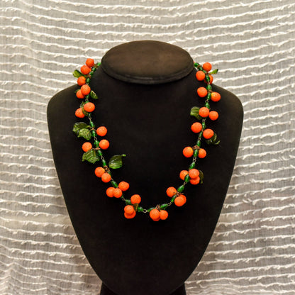 Venetian glass necklace with vibrant orange berry-shaped beads and green leaves, displayed on a black bust against a textured beige background.